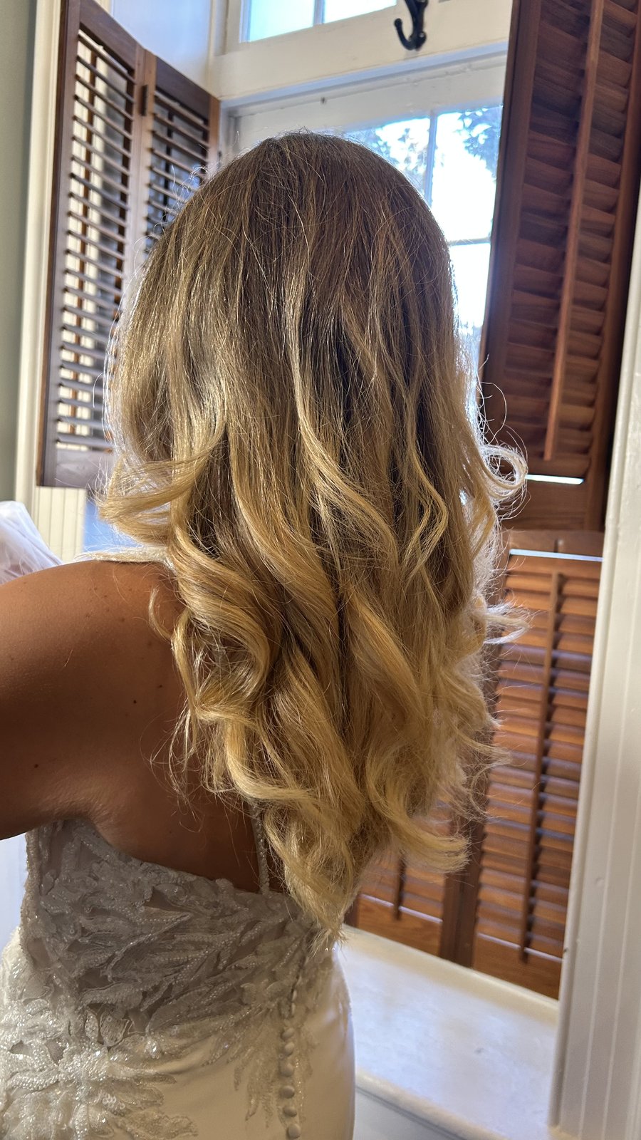 Long bridal balayage waves cascading down a white lace bodice, plantation shutters behind.